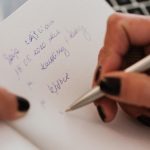 Crop anonymous female with manicured hands writing down notes in notebook with white sheets by using stylish silver pen near modern laptop